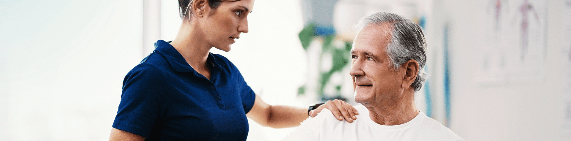 A healthcare worker, wearing a blue uniform, gently places her hand on an elderly man's shoulder and looks at him with concern, while the man, with gray hair and a white shirt, looks back at her with a calm expression.