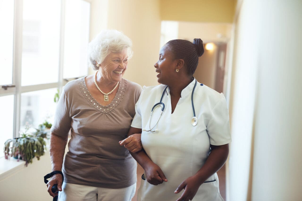A smiling elderly woman with white hair and a nurse with dark hair in a bun walk together in a bright hallway, engaging in conversation. The nurse wears a white uniform and has a stethoscope around her neck.