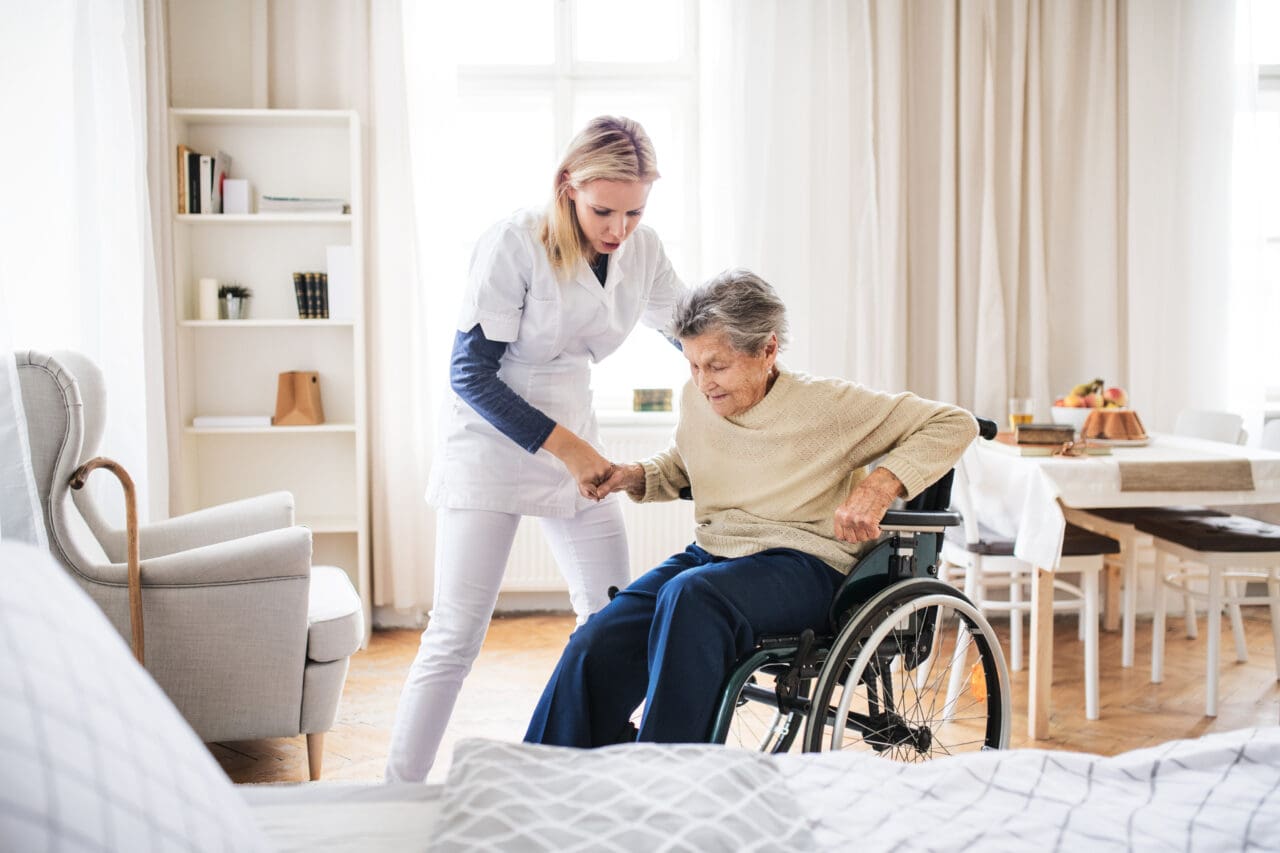 A caregiver assisting an elderly woman in a wheelchair, holding her hand and smiling in a bright, cozy room with large windows, a bookshelf, and a dining table with food in the background.