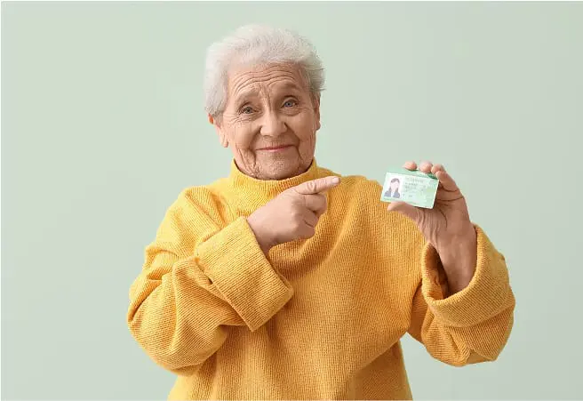 An elderly woman with short white hair is smiling and pointing to her driver’s license, which she is holding up in her right hand. She is wearing a yellow sweater and standing against a light green background.