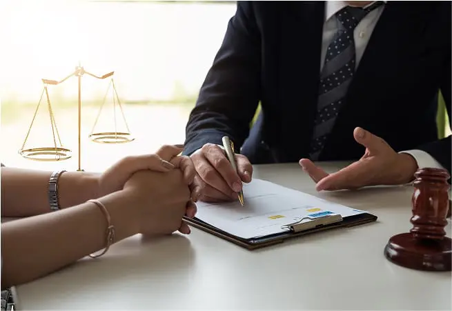 A person in a suit and tie is sitting at a table, signing a document with a pen, while another person holds their hands. A gavel and a set of balance scales are also on the table, indicating a legal or formal setting.