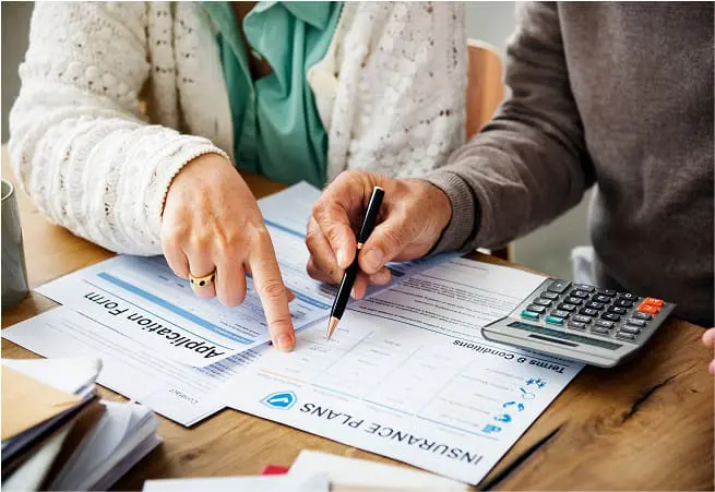 Two people are reviewing documents on a wooden table, with one pointing to a form labeled 