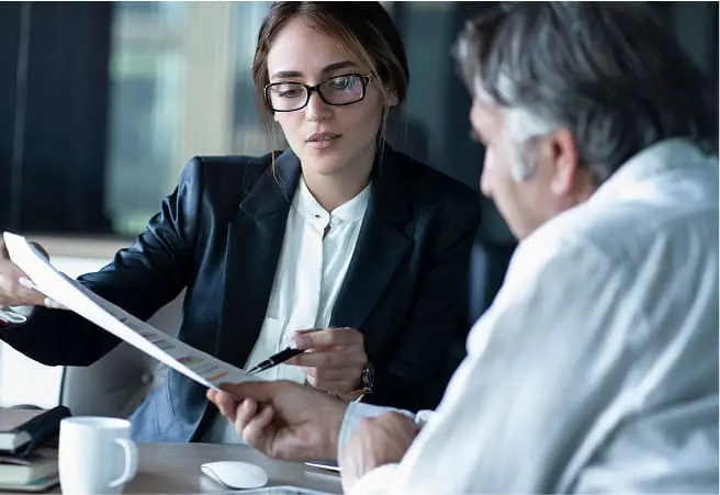 A young woman in glasses, dressed in a dark blazer and white shirt, sits at a table with an older man in a white shirt. They are reviewing documents together in a professional setting.
