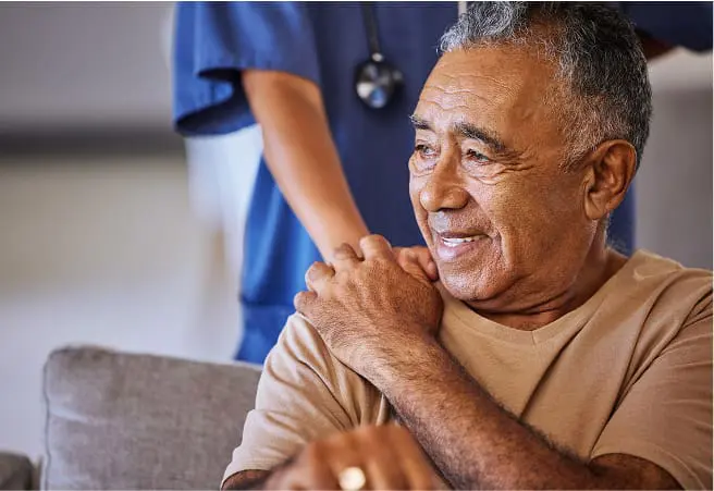 An elderly man sitting on a couch smiling, with a healthcare worker in blue scrubs standing behind him. The man has his right hand on his shoulder, and a stethoscope is visible around the worker's neck.