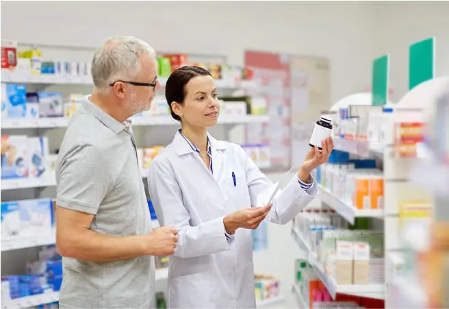 A pharmacist and an older male customer are standing in a pharmacy aisle; the pharmacist is holding a bottle of medication and showing it to the customer, who is attentively looking at it. Shelves stocked with various medicines and health products are in the background.