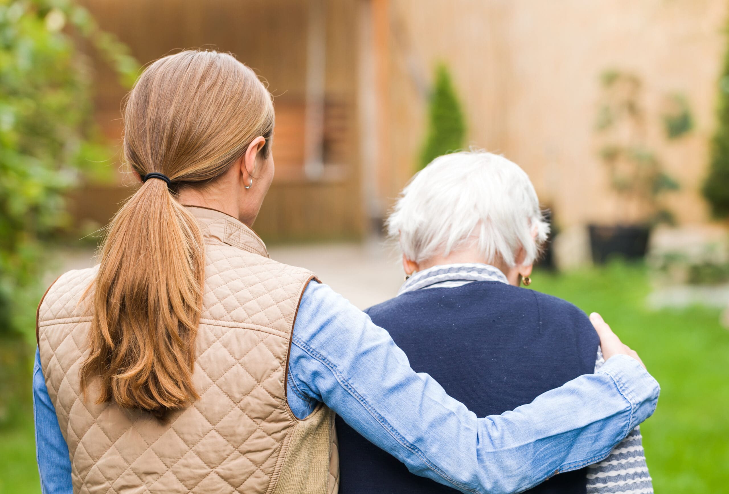 A young woman with long, light brown hair tied back in a ponytail, wearing a beige quilted vest over a blue shirt, has her arm around an elderly woman with short white hair, dressed in a dark sweater and a striped scarf. They are outdoors, standing close together in a garden or yard with green grass and blurred plants and buildings in the background.